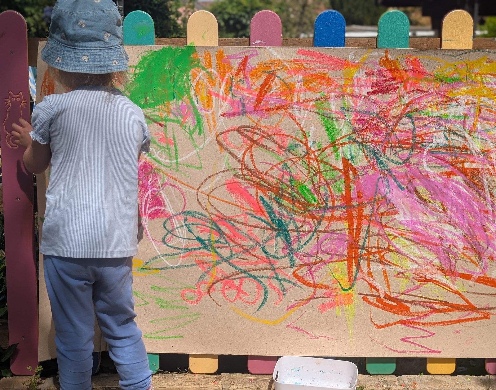 A child in a sun hat drawing at a large outdoor mark-making board, seen from behind