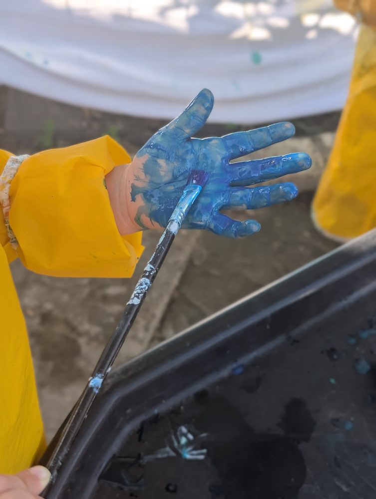 A child's open hand covered in blue paint, holding a paintbrush