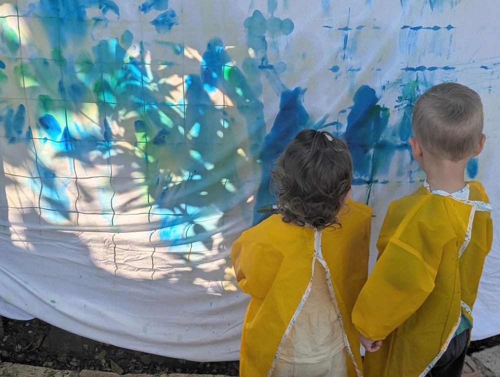 Two children in aprons painting a large fabric sheet in dappled sunlight, seen from behind